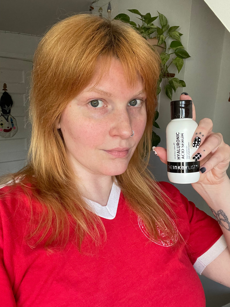 A woman with red hair holds up a small white bottle with a black cap, showing the brand name 'INKEY LIST'. She is wearing a red shirt in an indoor setting with a plant in the background.