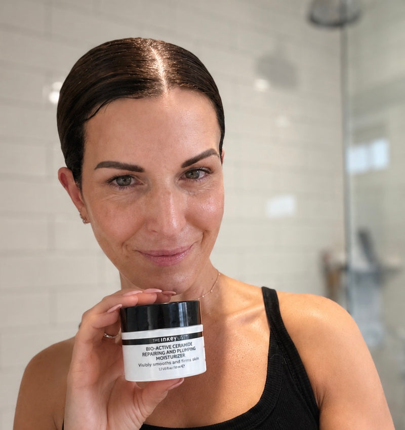 A woman in a black tank top holds a jar of Cream in a bright bathroom setting with white subway tiles.