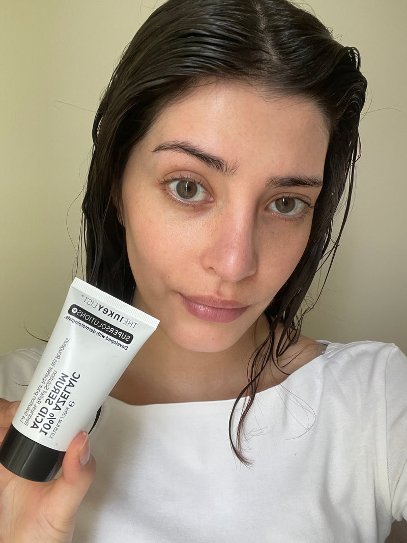 A woman with wet dark hair holds up a white tube of 10% Azelaic serum against a plain beige wall.