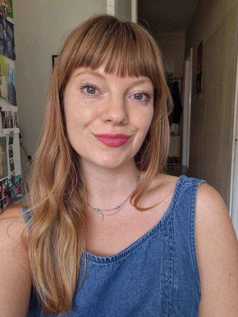 A woman with light brown hair and bangs wearing a blue denim tank top, photographed in a casual indoor setting.