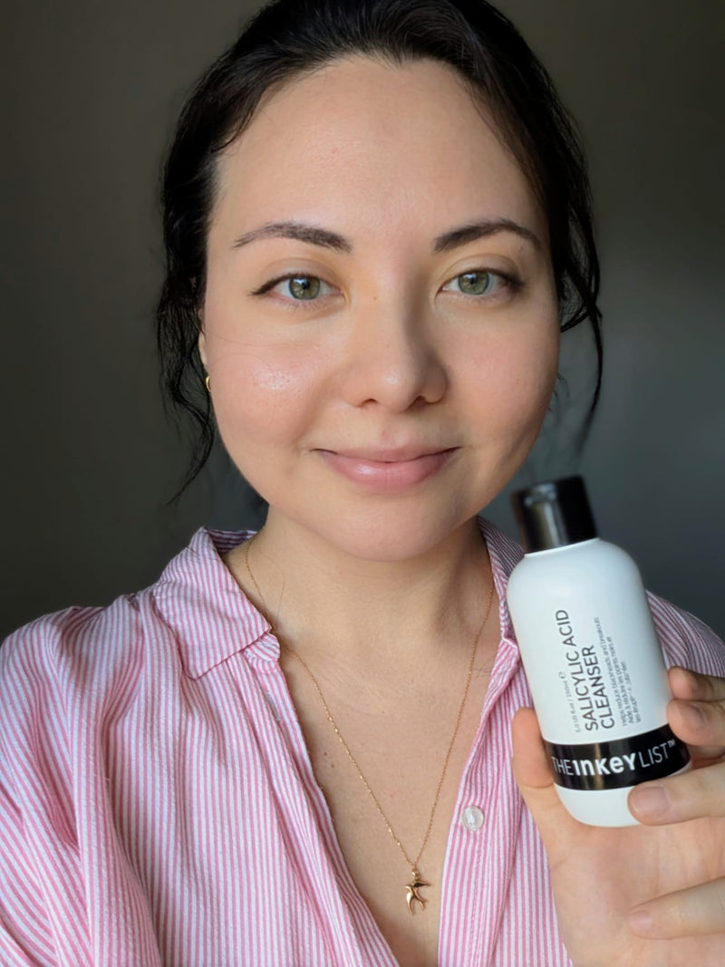 A woman with dark hair pulled back, wearing a pink and white striped button-up shirt, holding a white bottle of 'The Inkey List' Salicylic Acid Cleanser against a dark gray background.