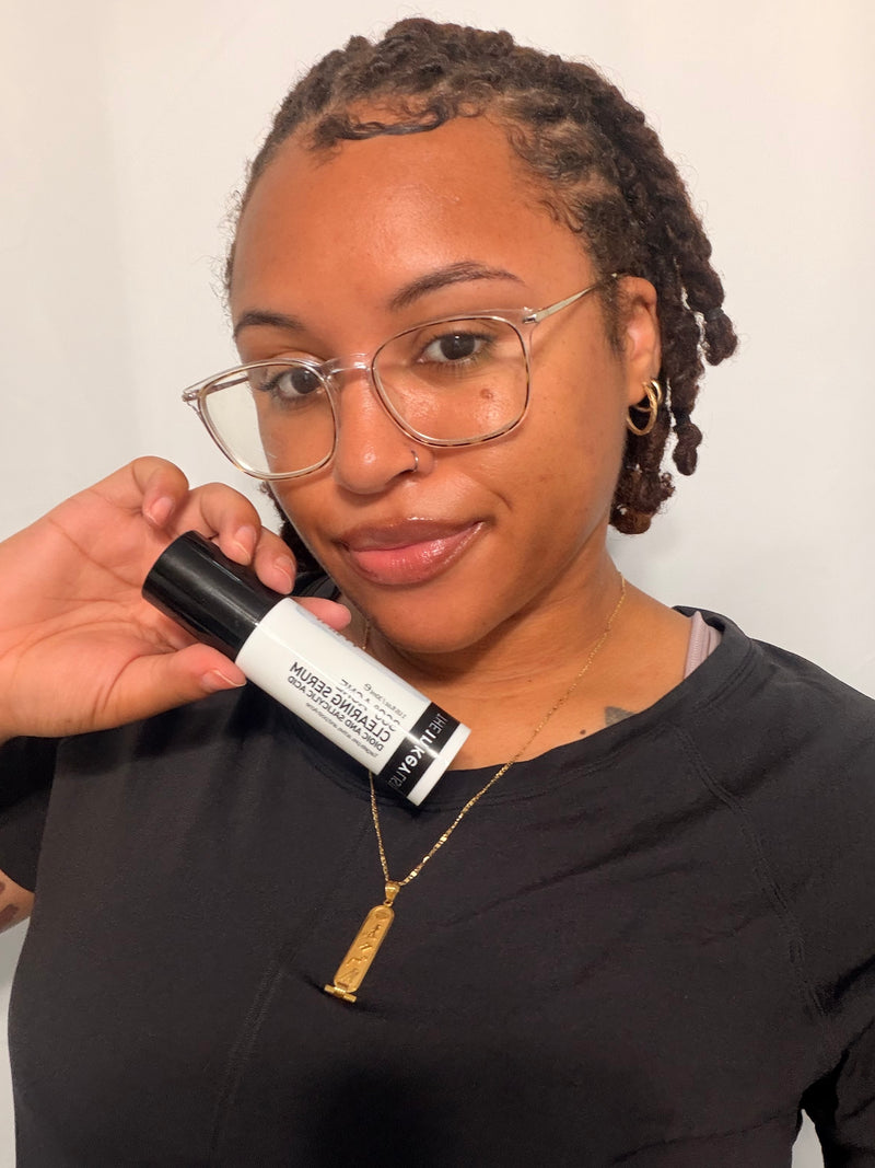 A woman with glasses and short hair holds a black and white bottle of '360 Serum' next to her face, wearing a black shirt and a gold pendant necklace against a plain light background.