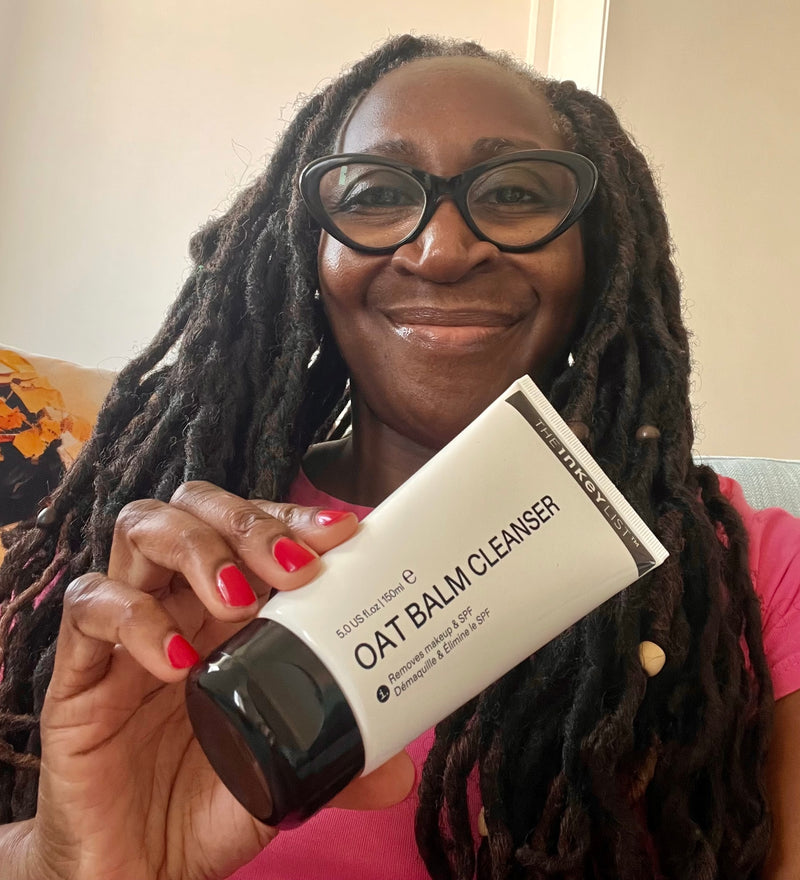 A smiling woman with long black hair and glasses holds up a white tube of 'The Inkey List' Oat Balm Cleanser. The product packaging is white with black text and a black cap, and the woman has red nail polish.