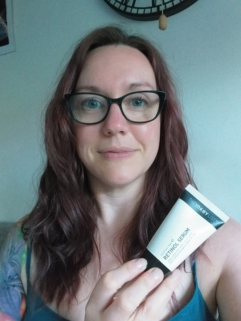 A woman with long reddish-brown hair and black-rimmed glasses holds a white tube of The Inkey List Retinol Serum in front of her face, set against a domestic background with a wall clock.