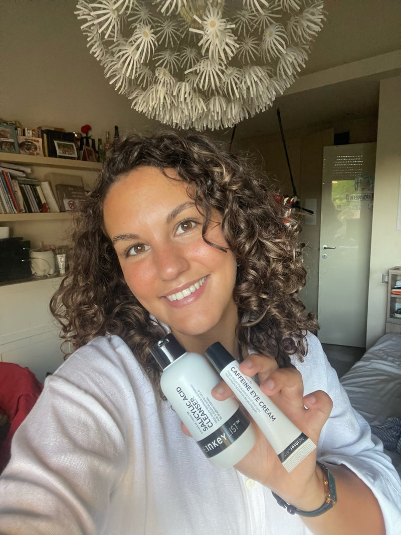 A smiling woman with curly brown hair holds up two skincare products, in a selfie-style photo taken in a bedroom with a decorative hanging light overhead.
