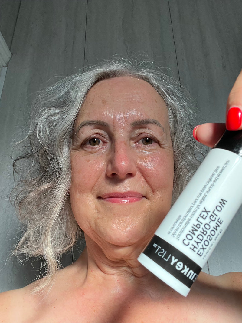 A close-up selfie of a woman with gray hair holding a white bottle of Exosome Complex against a gray paneled background.