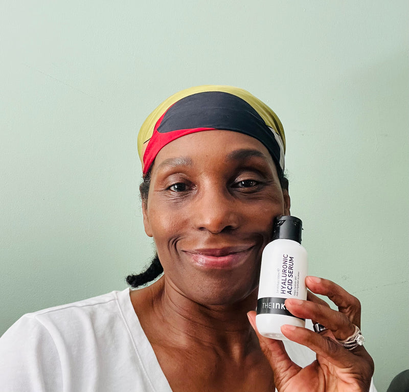A woman wearing a black, yellow, and red headband holds up a white bottle of The Inkey List Hyaluronic acid serum against a light mint green wall.