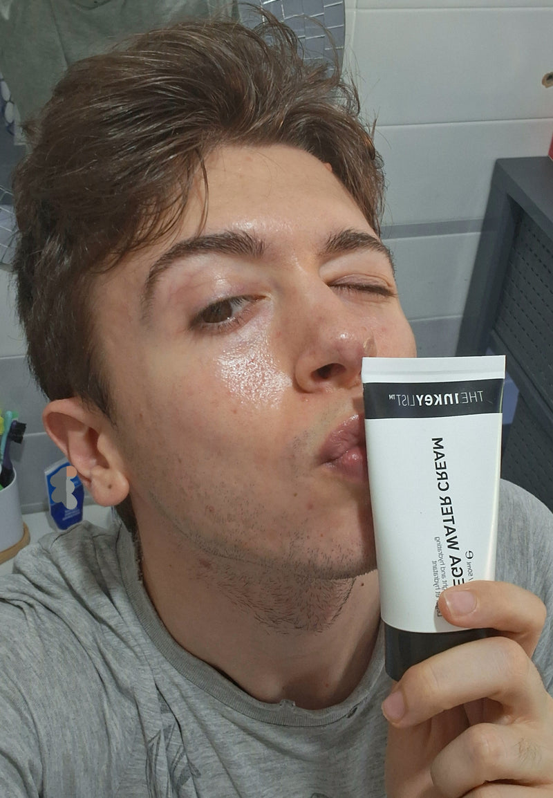 A man in a gray t-shirt holding a white and black tube of 'The Inkey List' Omega Water Cream to his lips in a bathroom setting.