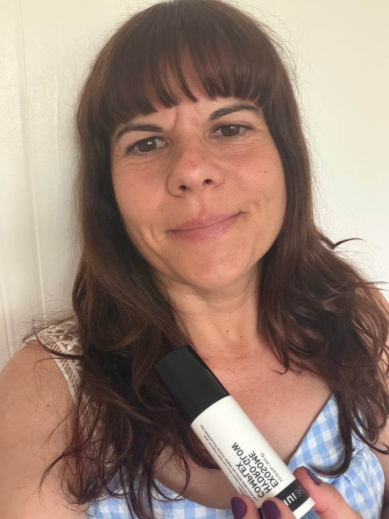 A woman with brown hair holding a white and black roll-on bottle of exosome serum against a plain off-white wall background.