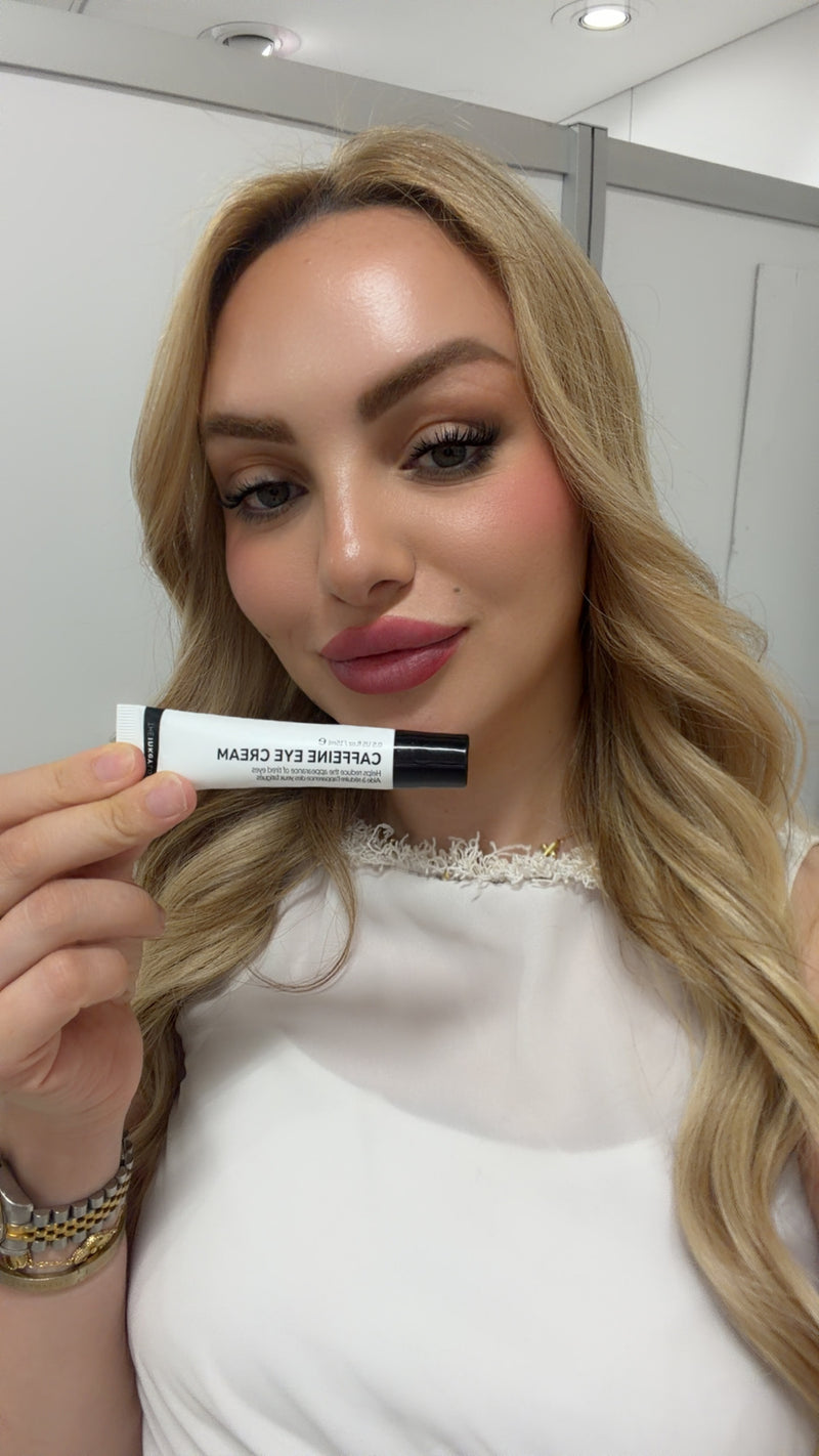 A woman with blonde hair holds a white tube of caffeine eye cream in front of her face. The tube is white with black text and black caps on both ends.