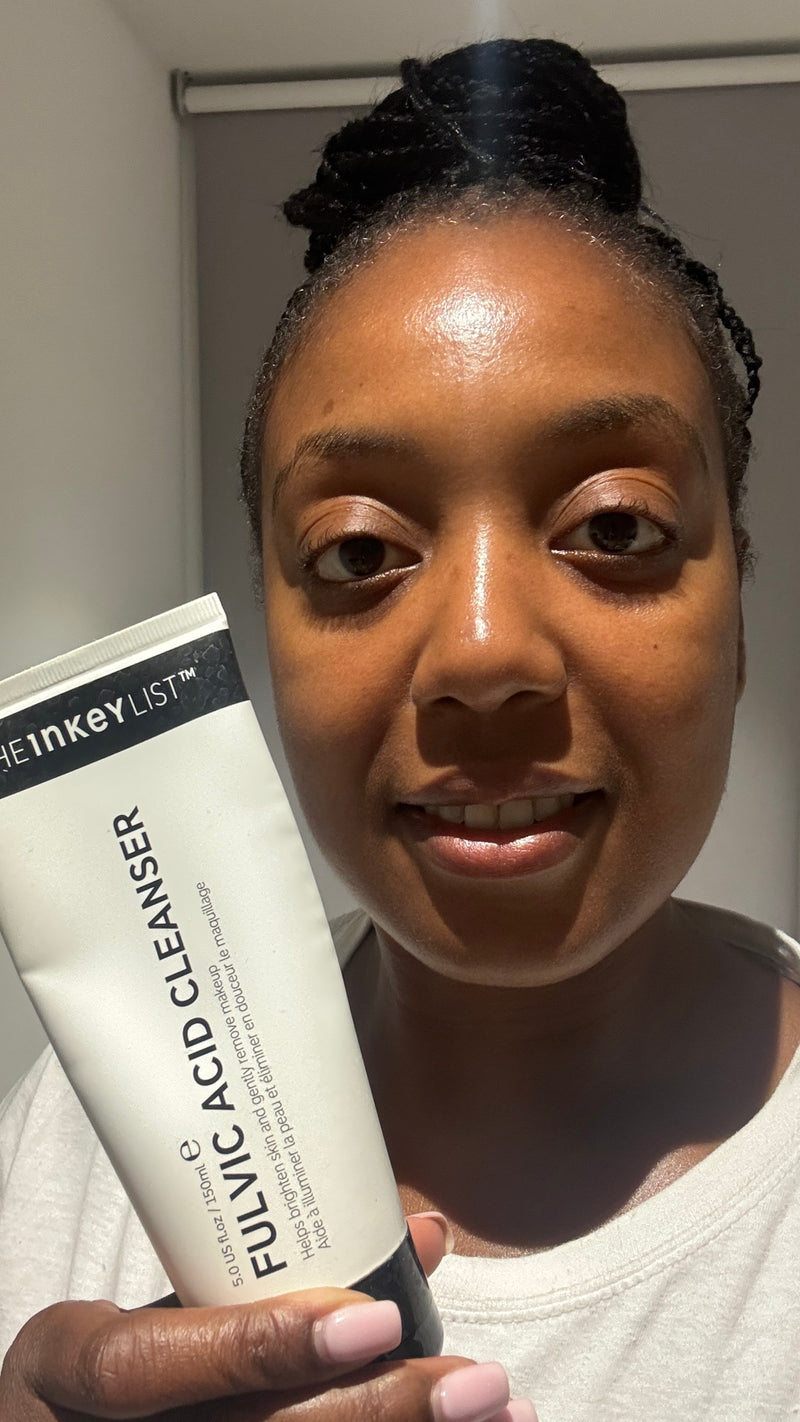 A woman holding a white tube of inkey List Fulvic Acid Cleanser in front of her face, set against a plain indoor wall.