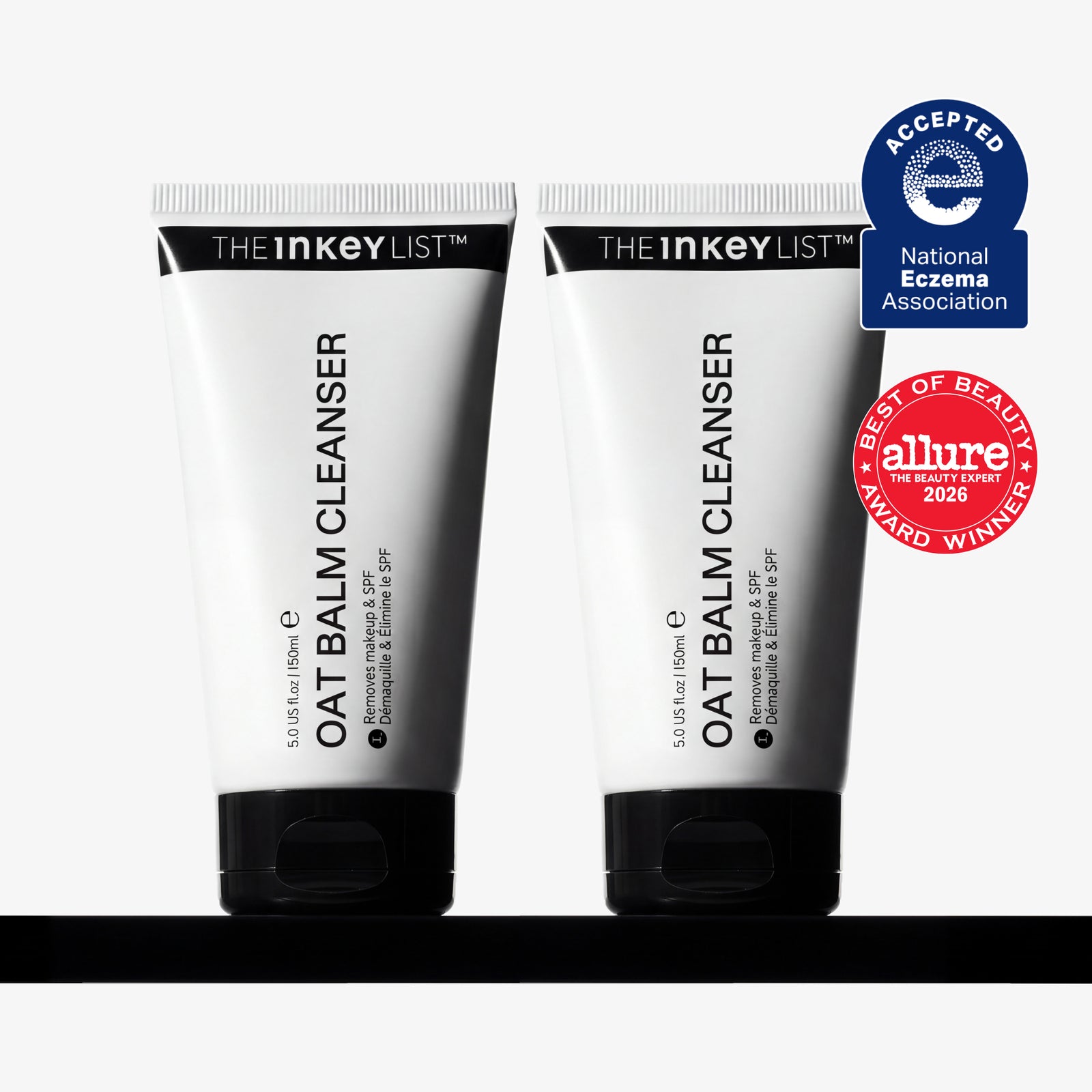 Two white squeeze tubes of THE INKEY LIST OAT Balm Cleanser with black caps, displayed side-by-side on a black surface against a white background, with two award badges on the right.