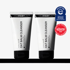 Two white squeeze tubes of THE INKEY LIST OAT Balm Cleanser with black caps, displayed side-by-side on a black surface against a white background, with two award badges on the right.