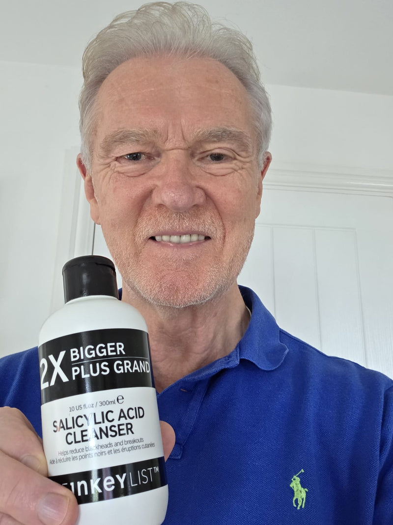 An older man with gray hair and a beard wearing a blue polo shirt with a green embroidered polo player logo, holding up a white bottle of '2X Bigger Plus Grand Salicylic Acid Cleanser' by 'INKEY'.