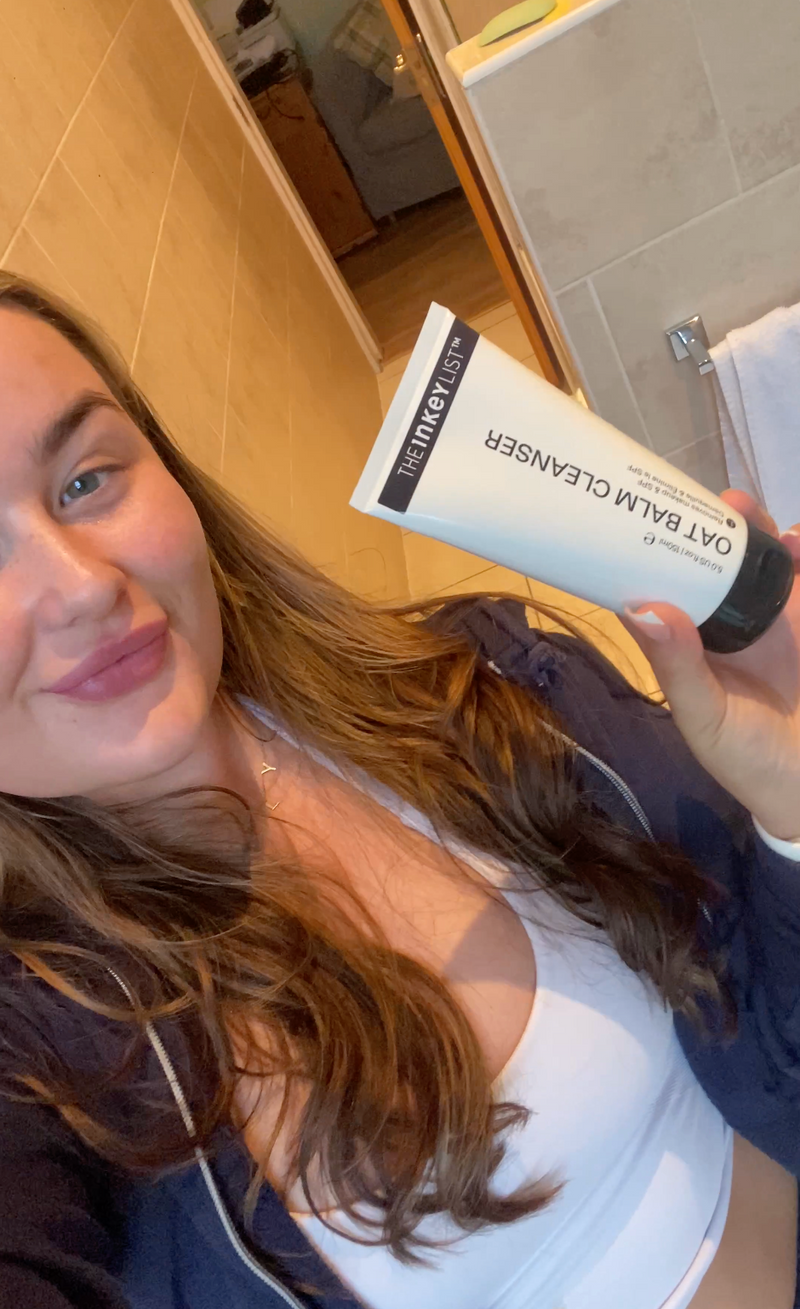 A woman holding a white tube of Oat Balm Cleanser in a bathroom setting.