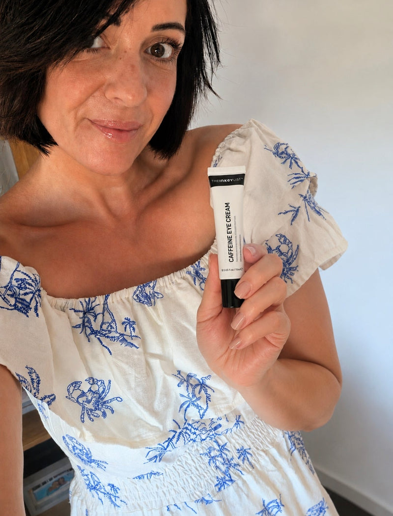 A woman holding a black and white tube of Caffeine eye cream wearing a white off-the-shoulder dress with a blue floral pattern.