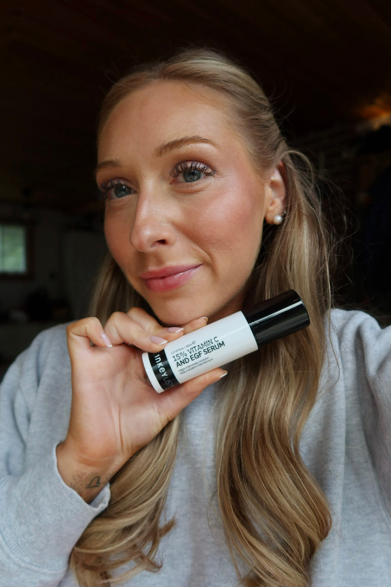 A woman with blonde hair holding a white and black tube of 15% Vitamin C Serum, wearing a light gray sweatshirt in an indoor setting.