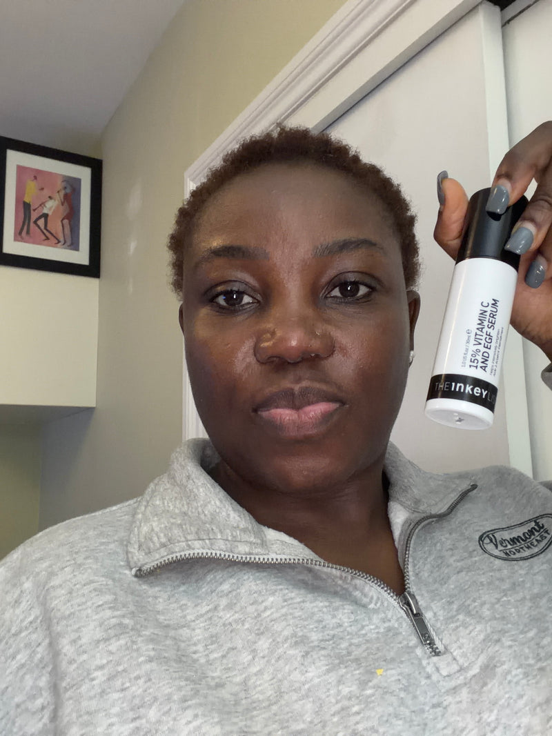A woman holding a white and black The Inkey List 15% Vitamin C Serum bottle in front of her face, wearing a gray zip-up sweatshirt in an indoor setting.