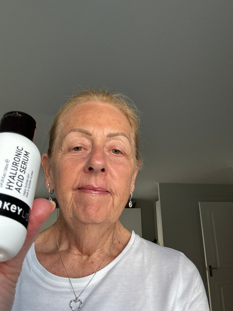 An older woman holding up a white bottle of Hyaluronic Acid Serum in front of her face, wearing a white t-shirt and silver jewelry in an indoor setting.